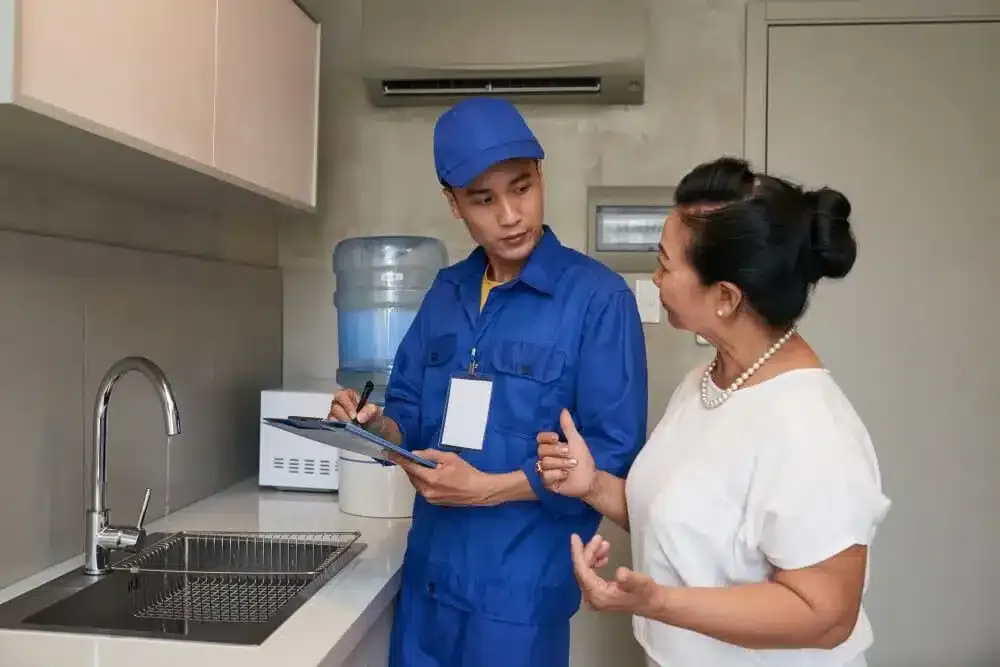 Asian-male-plumber with uniform-talking-senior-female-homeowner-kitchen