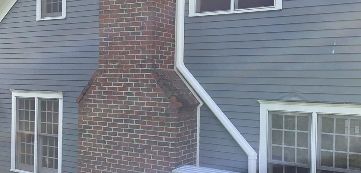 Bosch heat pump with a white line-set cover installed beside a brick chimney on a colonial home